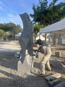 Escultor a trabalhar bloco de granito cinzento de grão fino; poeira de corte e ferramentas visíveis sobre brita.