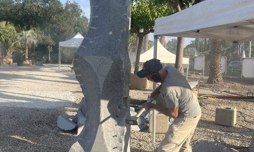 Escultor a trabalhar bloco de granito cinza de grão fino; poeira de corte e ferramentas visíveis sobre brita.