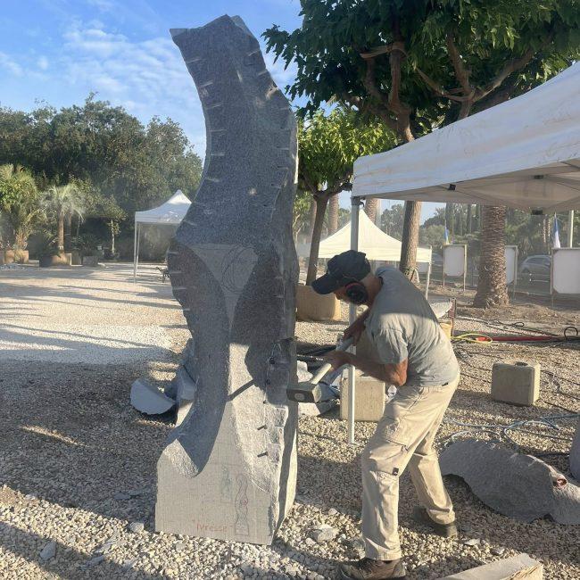 Escultor a trabalhar bloco de granito cinza de grão fino; poeira de corte e ferramentas visíveis sobre brita.